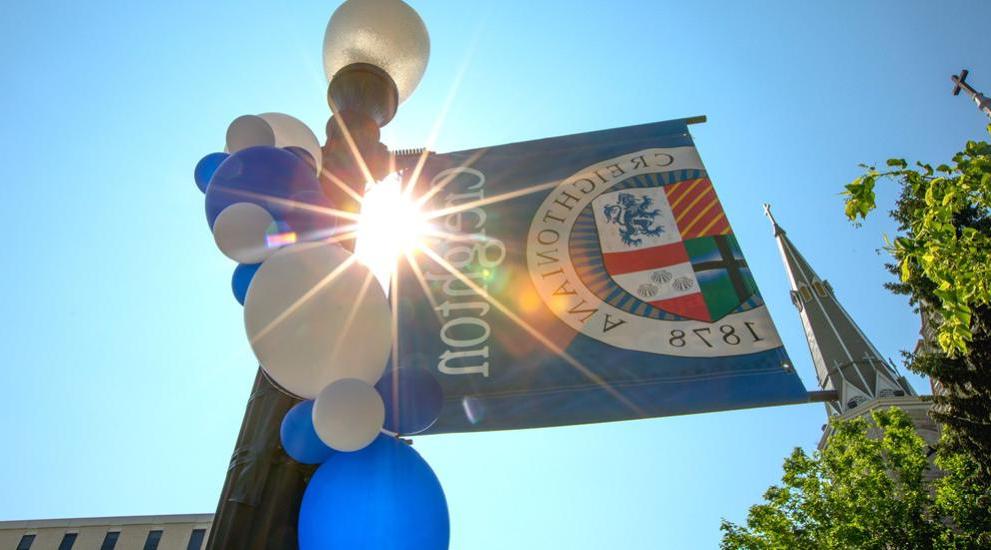 Sun burst coming through Creighton flag on pole ordained with blue and white blues.